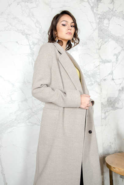 Beauty Fashion Model Girl with curls in light beige coat in a bright white loft style studio. Beautiful Luxury Winter Woman. Beauty and fashion. Spring and autumn clothes fashion Girl posing at studio - Φωτογραφία, εικόνα