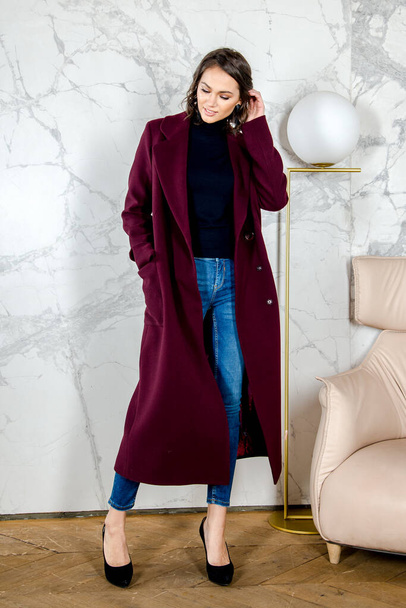 Beauty Fashion Model Girl with curls in burgundy coat in a bright white loft style studio. Beautiful Luxury Winter Woman. Beauty and fashion. Spring and autumn clothes fashion. Girl posing at studio. - Foto, Imagen
