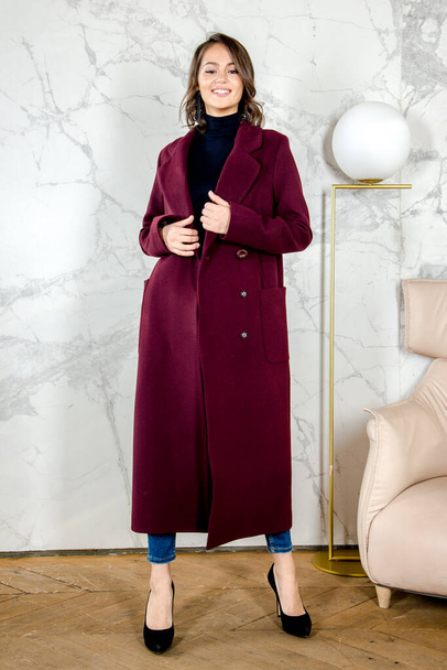 Beauty Fashion Model Girl with curls in burgundy coat in a bright white loft style studio. Beautiful Luxury Winter Woman. Beauty and fashion. Spring and autumn clothes fashion. Girl posing at studio. - 写真・画像