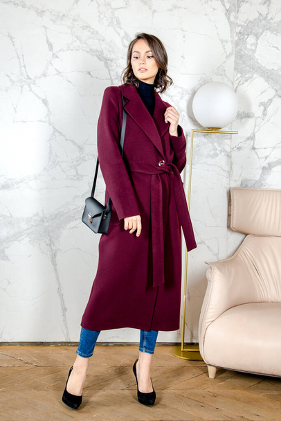 Beauty Fashion Model Girl with curls in burgundy coat in a bright white loft style studio. Beautiful Luxury Winter Woman. Beauty and fashion. Spring and autumn clothes fashion. Girl posing at studio. - Фото, изображение