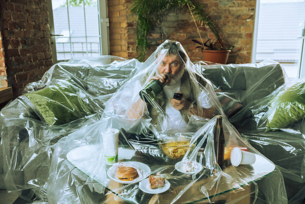 Senior man covered with plastic, eating fast food and drinking beer - environmental pollution by people concept - 写真・画像