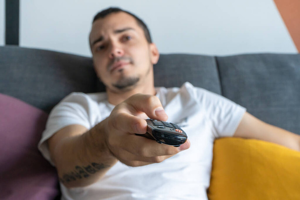A thin guy in home clothes sits on a sofa and watches TV. Bored guy with tv remote control. - Foto, Imagen