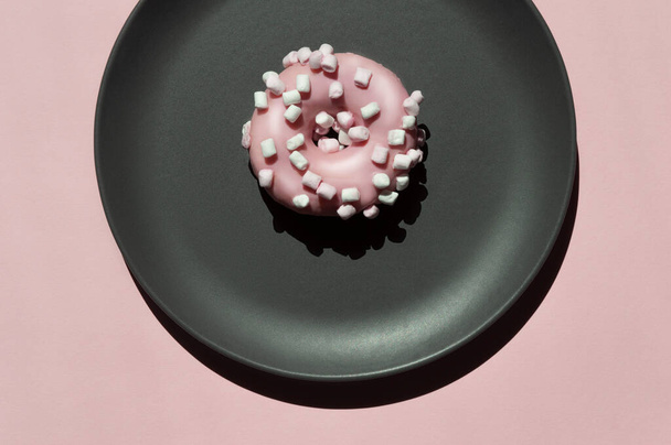 One pink donat with white marshmallows on the dark grey ceramic plate on the light coral background. Geometric sunny creative trendy composition with shadows. Art idea for celebration donat day - Photo, Image