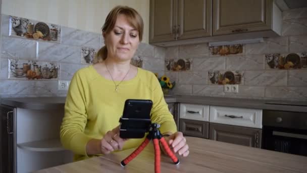 Молодая кавказская улыбающаяся женщина со смартфоном общается в онлайновом чате и социальных сетях, находясь дома на кухне. Блогер ведет прямые трансляции для своих последователей. Досуг
. - Кадры, видео
