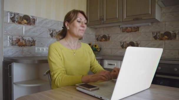 Young caucasian smiling woman waves her hand in greeting during online communication using a laptop, sitting at home at a table in the kitchen. Social networks and home leisure. - Filmagem, Vídeo