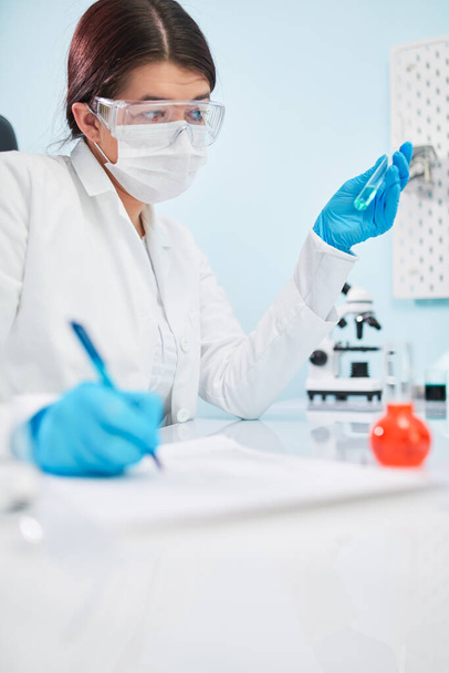 Woman lab technician with test tube in hand makes note. Coronavirus epidemic - Zdjęcie, obraz