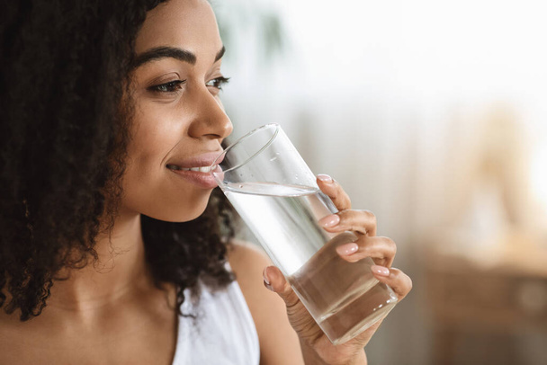 Здоровая жидкость. Улыбающаяся чернокожая женщина пьет воду из стекла и смотрит в сторону
 - Фото, изображение
