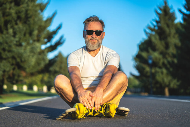 Starý muž ve slunečních brýlích s kolečkovými bruslemi sedícími na asfaltu - Fotografie, Obrázek