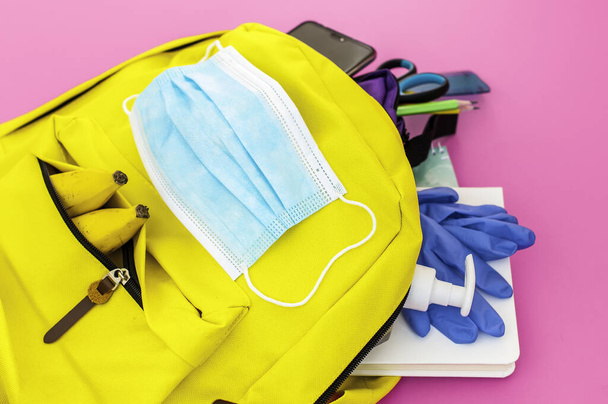 Yellow backpack full of school must-have tools also with face mask, hydro alcoholic gel and latex gloves to protect from covid-19 on the pink background - Zdjęcie, obraz