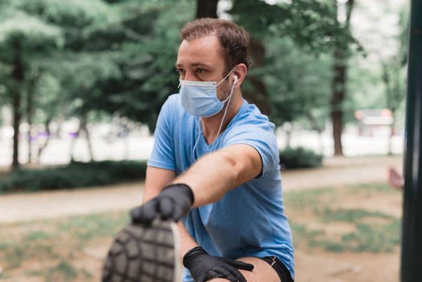 医療用マスクと手袋、スマートフォンとイヤホンが動作し、都会の環境でジョギングを持つスポーツマン. - 写真・画像
