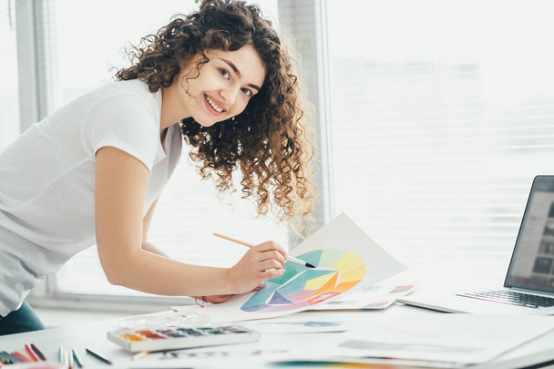 Кудрявая девушка с кистью рисует картину за столом
 - Фото, изображение