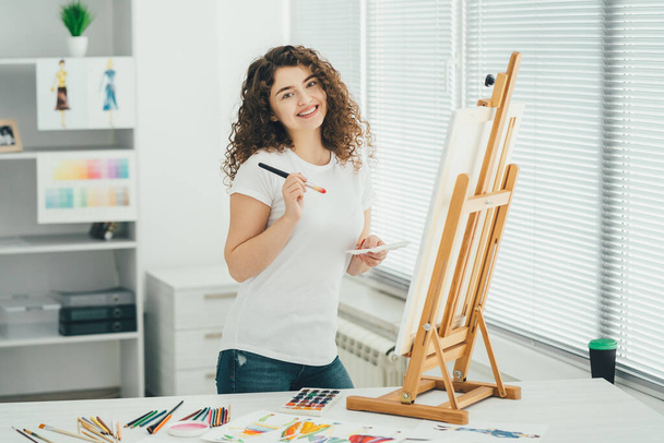 The cute woman with an art brush painting a picture on the easel - Φωτογραφία, εικόνα