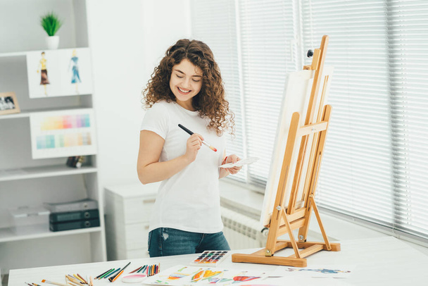 The cute woman with an art brush painting a picture on the easel - Фото, изображение