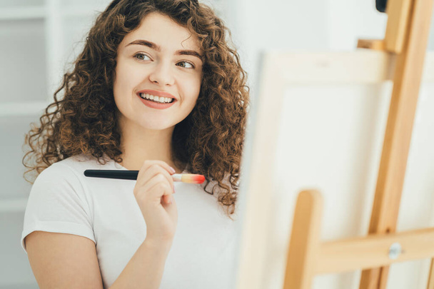 Kudrnatá žena s uměleckým štětcem maluje obrázek na stojan - Fotografie, Obrázek