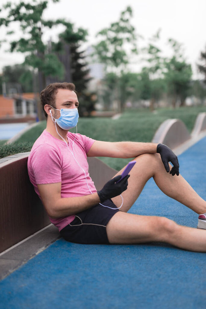 医療用マスクと手袋、スマートフォンとイヤホンが動作し、都会の環境でジョギングを持つスポーツマン.  - 写真・画像