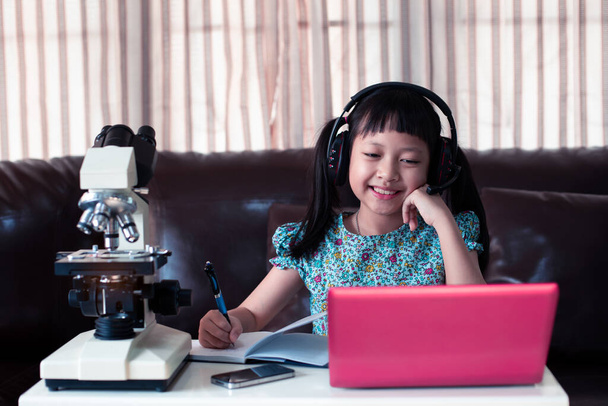 Ασιατικό κοριτσάκι φορώντας ακουστικά μάθησης σε απευθείας σύνδεση με τη χρήση φορητού υπολογιστή και μικροσκοπίου στο σπίτι, έννοια Online εκπαίδευση, Κοινωνική απόσταση κατά τη διάρκεια της καραντίνας - Φωτογραφία, εικόνα