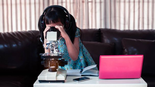 Ασιατικό κοριτσάκι φορώντας ακουστικά μάθησης σε απευθείας σύνδεση με τη χρήση φορητού υπολογιστή και κοιτάζοντας μικροσκόπιο στο σπίτι, Online εκπαίδευση έννοια, Κοινωνική απόσταση κατά τη διάρκεια της καραντίνας - Φωτογραφία, εικόνα
