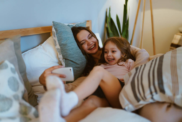 mom and daughter in the morning in bed laugh and take a selfie - Фото, изображение