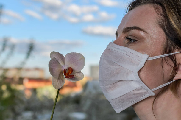 Young girl in medical protective mask sniffs an orchid flower - Фото, изображение