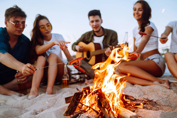 Group of young friends sitting on beach and fry sausages. One man is playing guitar. Summer holidays, vacation, relax and lifestyle consept. Camping time. - 写真・画像