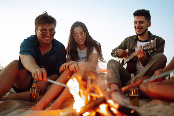 Group of young friends sitting on beach and fry sausages. One man is playing guitar. Summer holidays, vacation, relax and lifestyle consept. Camping time. - Foto, afbeelding