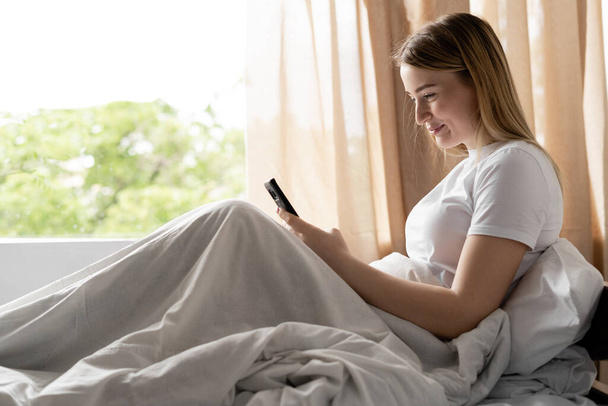 A young woman sits on a bed early in the morning. Reads great news on the phone. Cute smile on face. Chatting with friends online. Write a message. Window and place for text. - Foto, Imagem