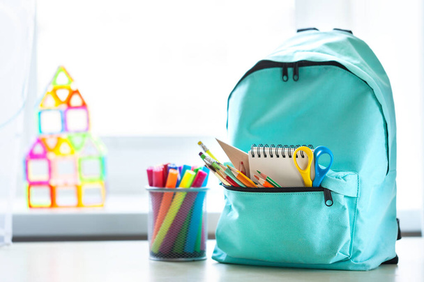 Blue school backpack with school supplies on table in children room across from the window in sunny day. Back to school concept. - Zdjęcie, obraz