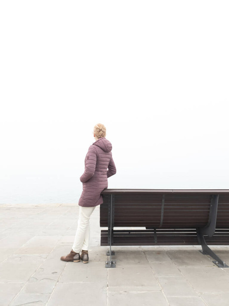 Woman in despair sitting on a lonely bench and looking at grey distant horizon on a ocean foggy day. - Fotó, kép