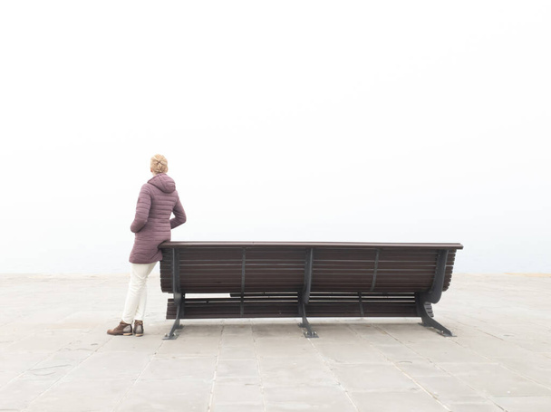 Жінка в розпачі сидить на самотній лавці і дивиться на сірий далекий горизонт на океанський туманний день
. - Фото, зображення