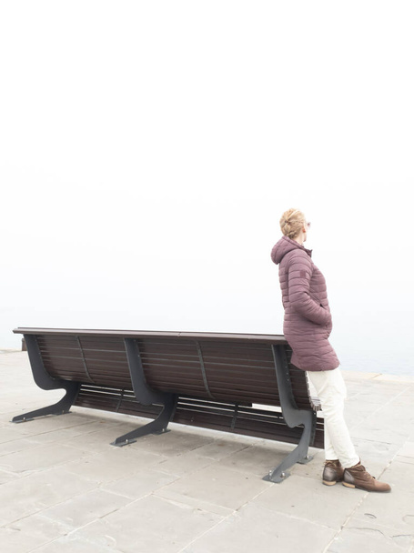 vrouw in wanhoop zittend op een eenzame bank en kijkend naar grijze verre horizon op een oceaan mistige dag. - Foto, afbeelding