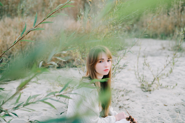 portrét mladé dívky v bílých šatech, teplý pletený svetr a černé boty mezi suchými rákosím a zelenými keři na břehu řeky s bílým pískem - Fotografie, Obrázek