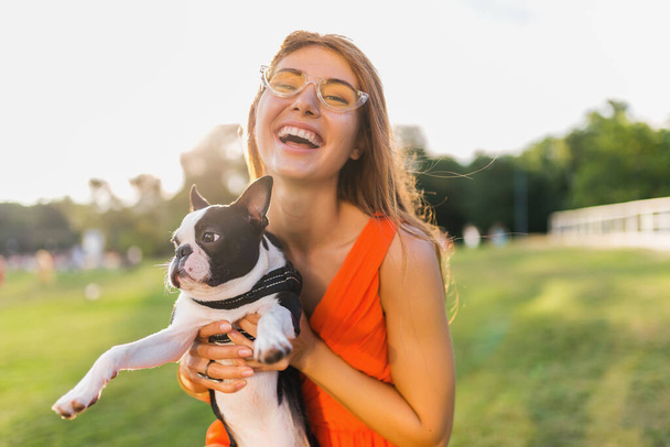 happy pretty woman park holding boston terrier dog, smiling positive mood, trendy summer style, wearing orange dress, sunglasses, playing with pet, having fun, sunny weekend entertainment - Foto, Imagem