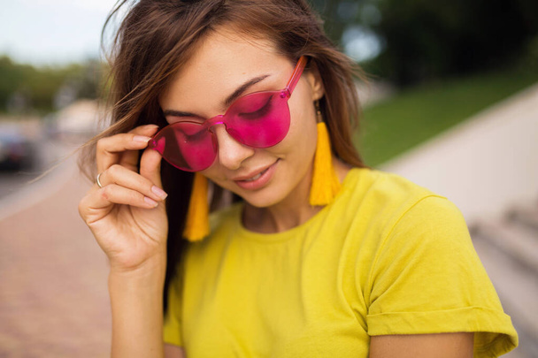 close up portrait of young attractive smiling woman having fun in city park, positive, happy, wearing yellow top, earrings, pink sunglasses, summer style fashion trend, stylish accessories, colorful - Фото, изображение
