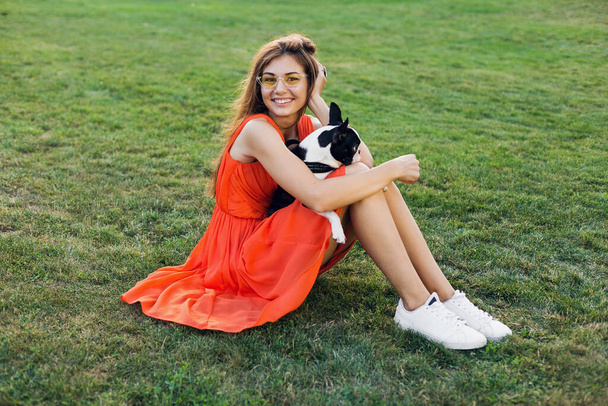 happy pretty woman sitting on grass in park, holding boston terrier dog, smiling positive mood, wearing orange dress, trendy style, slim legs, sneakers, playing with pet, summer fashion trend - Foto, afbeelding