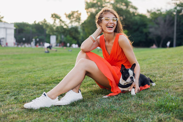 happy pretty woman sitting on grass in summer park, holding boston terrier dog, smiling positive mood, wearing orange dress, trendy style, slim legs, sneakers, playing with pet - 写真・画像