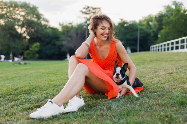 happy pretty woman sitting on grass in summer park, holding boston terrier dog, smiling positive mood, wearing orange dress, trendy style, slim legs, sneakers, playing with pet - Photo, Image