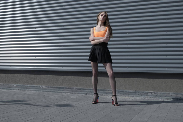 A young beautiful brunette in full growth against the background of a black building looks directly at the camera, dressed in a bright orange top, black fishnet tights and a black leather jacket - Valokuva, kuva