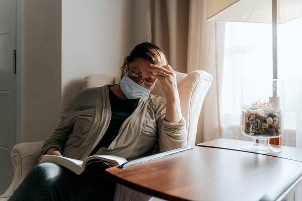 An older woman is sitting on the sofa with a book on her legs. She is worried about the pandemic. She is sick and she is wearing a protective mask. She is protecting her family. Pandemic concept. - Photo, Image