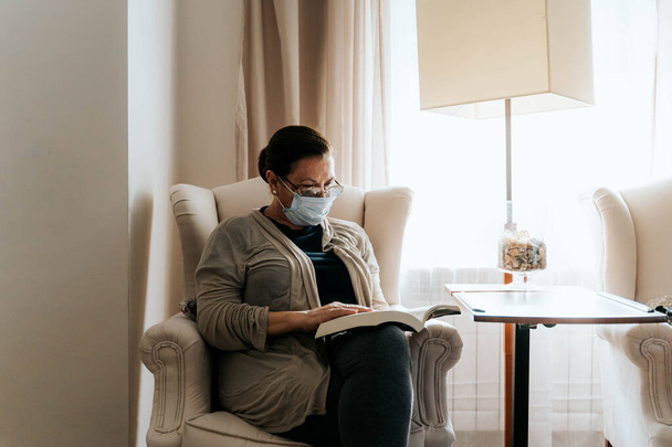 Starsza kobieta siedzi na kanapie czytając książkę. Jest chora i nosi maskę ochronną. Chroni swoją rodzinę i siebie. Koncepcja pandemii. - Zdjęcie, obraz
