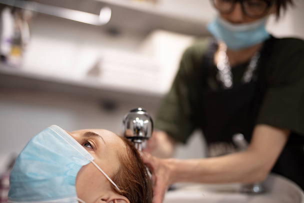 Κομμώτρια και πελάτισσα σε κομμωτήριο με ιατρικές μάσκες κατά τη διάρκεια πανδημίας ιών. Εργασία με μάσκα ασφαλείας. - Φωτογραφία, εικόνα