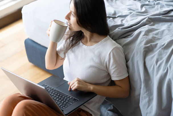 Beautiful young brunette girl working on a laptop and drinking coffee, sitting on the floor near the bed. Stylish modern interior. A cozy workplace. Shopping on the Internet. - Fotoğraf, Görsel