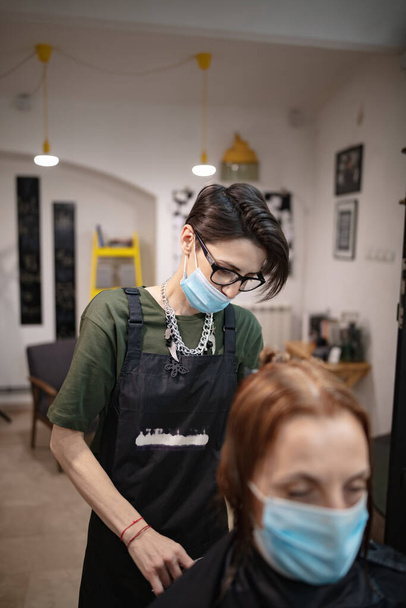 Peluquería y cliente en un salón con máscaras médicas durante la pandemia de virus. Trabajar con máscara de seguridad. - Foto, Imagen