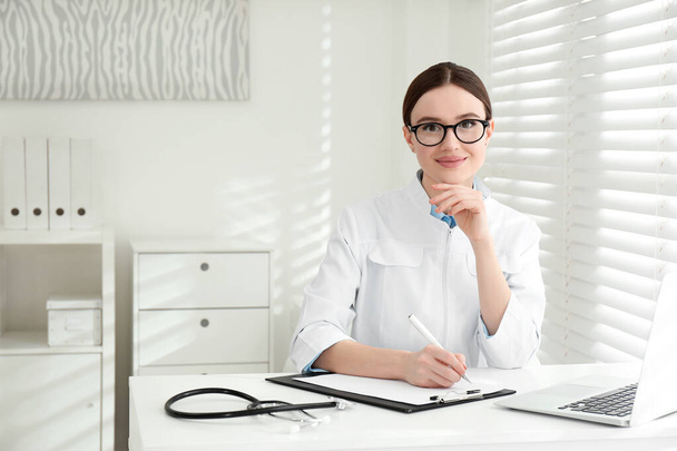Portrait of young female doctor in white coat at workplace - Fotoğraf, Görsel