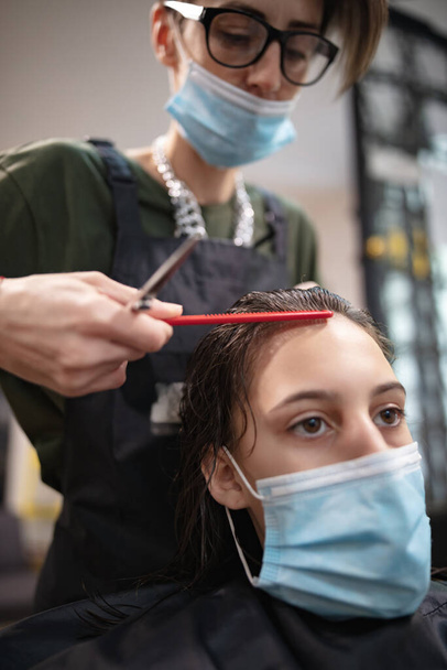 Парикмахер и девочка-клиент в салоне с медицинскими масками во время вирусной пандемии. Работа с защитной маской. - Фото, изображение
