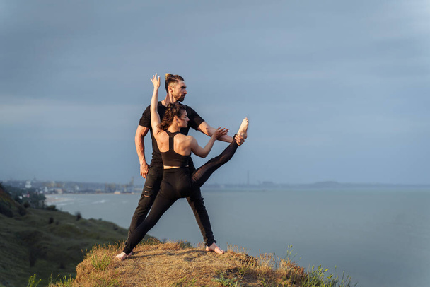 Akroyoga in nature. Fresh air, free space, untouched by human nature outside the city in the sun set. Talented athletes doing stretching poses. Sportsman holding girl's leg, they look at the sea. - Photo, Image