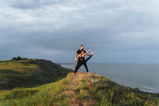 Akroyoga in nature. Fresh air, free space, untouched by human nature outside the city in the sun set. Talented athletes doing stretching poses. Sportsman holding girl's leg, they look at the sea. - Photo, Image