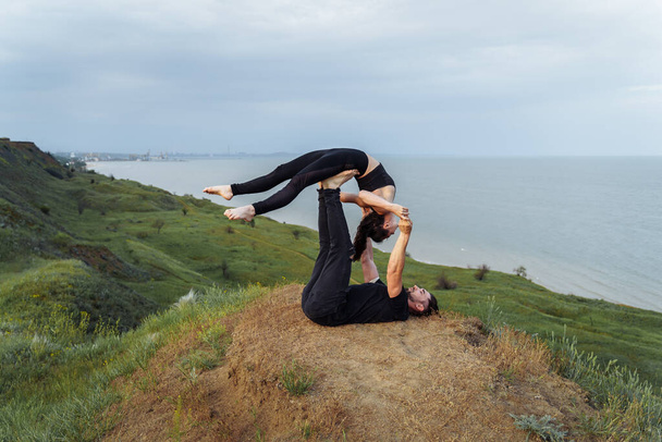 Koncepcja jogi akro. Para młodych sportowców ćwiczących parę lekcji jogi, mężczyzna i kobieta ćwiczący, równowaga ramion pozują na wzgórzu przeciwko morzu. Dziewczyna wygięła plecy, leżąc na nogach ahtlete - Zdjęcie, obraz