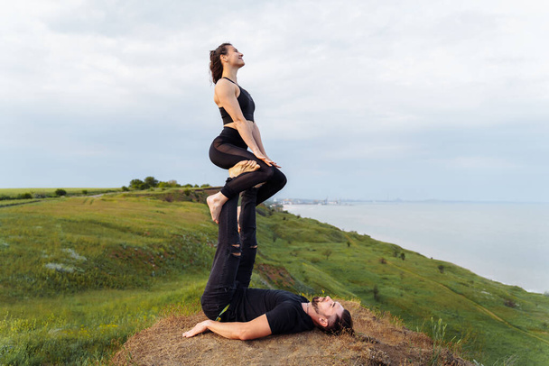 Acroyoga posture straddle throne. An athletic couple of sportsmen doing stunts on a cliff outside the city. The guy lying in the grass with his back down, and the girl sitting on his outstretched legs - Photo, Image
