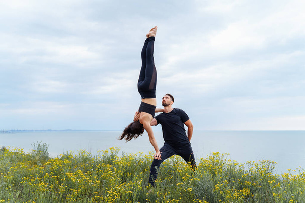 Two sportsmen practicing acro yoga together outdoors in the evening. Balancing girl legs up. Perfect flexibility. Athlete standing upside down. Holding a girl with one hand. Long shot - Photo, Image