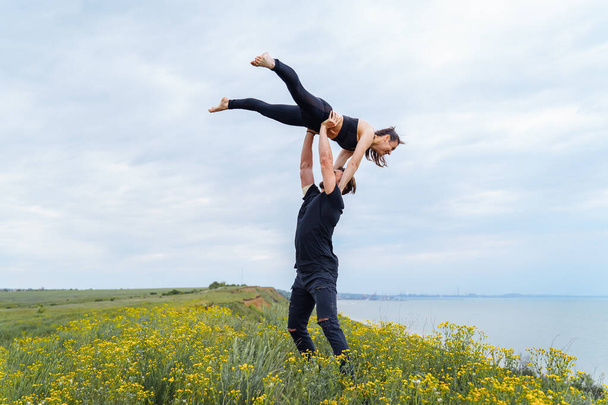 Lifestyle acroyoga and yoga poses. Pair of sportsman and sportswoman standing in the position of asana. Acrobat holding girl arched high back on the outstretched arms. Outdoors isolated from people - Photo, Image